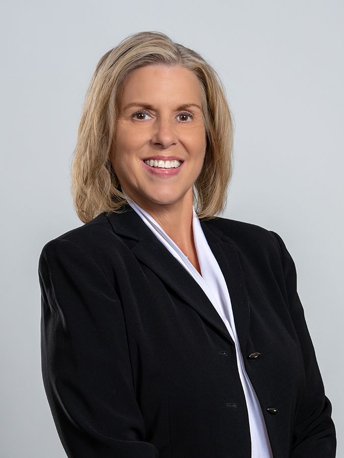 A person with blonde hair smiling while wearing a black blazer and white collared shirt against a gray background.