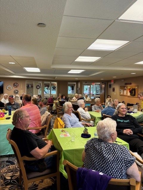 People sit at tables with green cloths in a well-lit indoor room, likely attending a group social gathering.