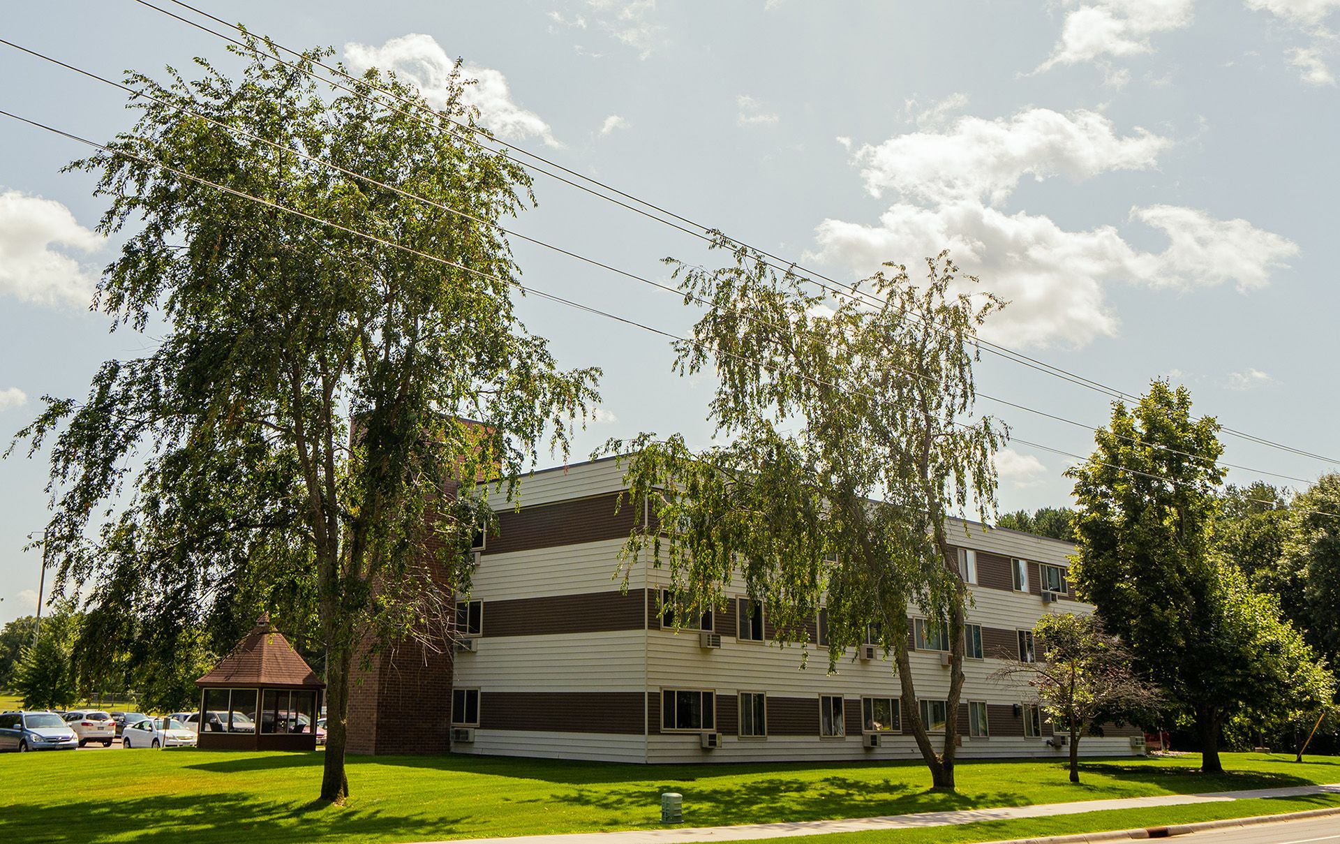 A three-story apartment building with horizontal brown and white siding stands on a grassy lawn with trees and a gazebo.