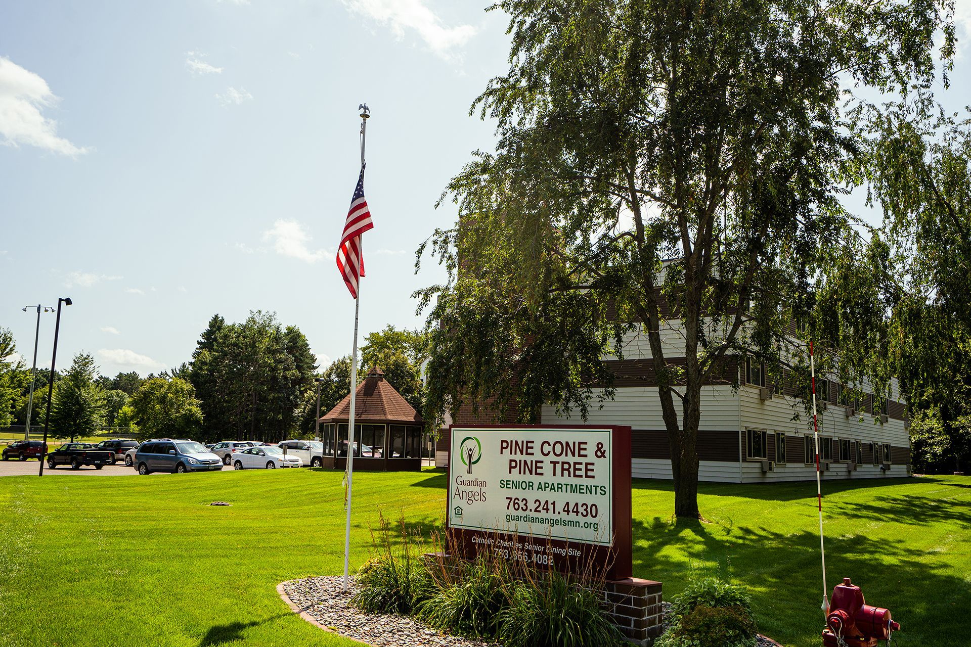 A sign for Pine Cone & Pine Tree resort in a sunny green yard with an American flag, trees, and parked cars nearby.