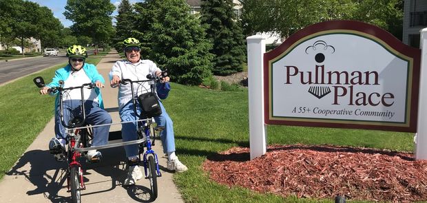 Two people in helmets ride a side-by-side tricycle past a Pullman Place sign on a sunny day.