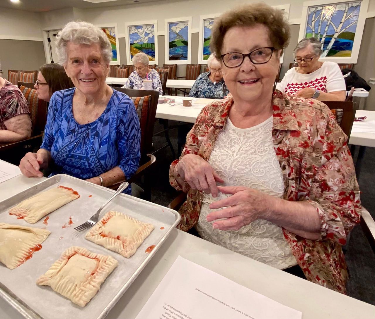Two people sitting at a table with a tray of prepared pastries, smiling toward the camera in a bright room.