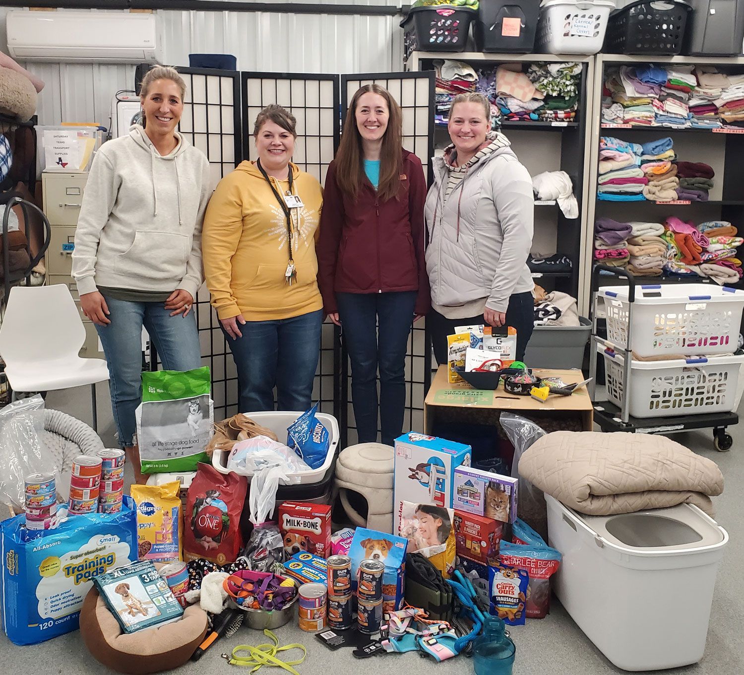 Four people stand in an animal supply donation center, surrounded by pet food, toys, and pet bedding on the floor.