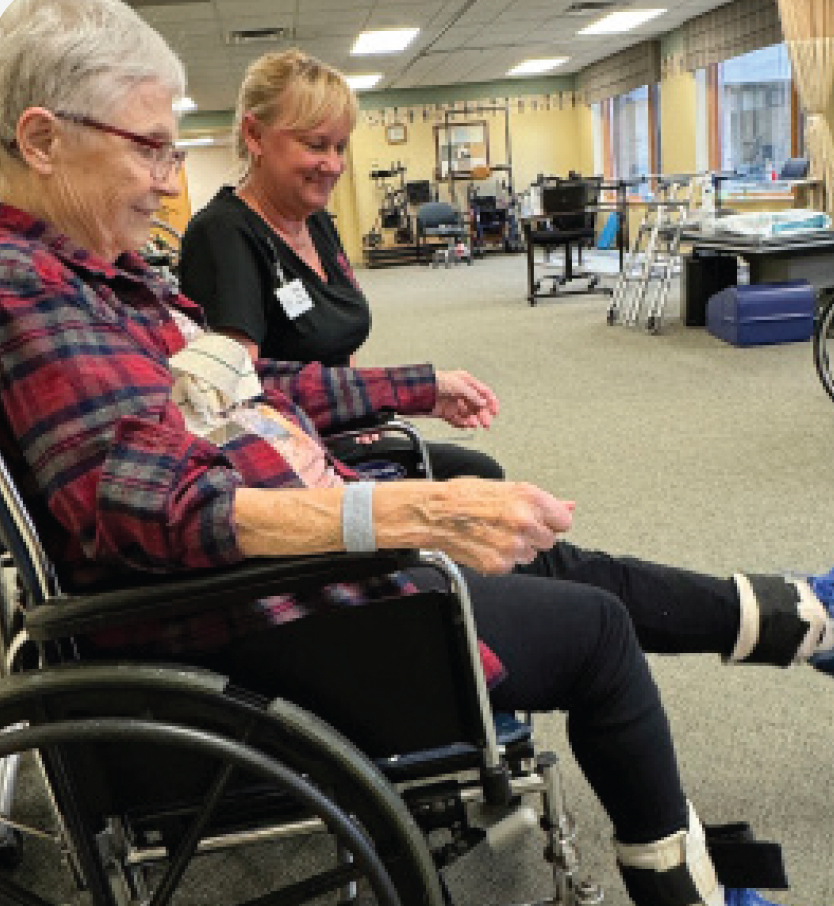 A person in a wheelchair performs a leg extension exercise with ankle weights, guided by a staff member in a therapy gym.