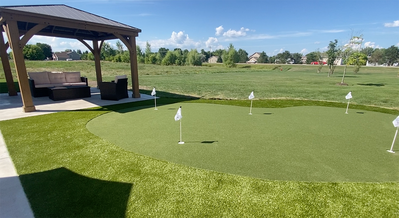 A backyard putting green with several flags sits next to a wooden gazebo with outdoor patio furniture.