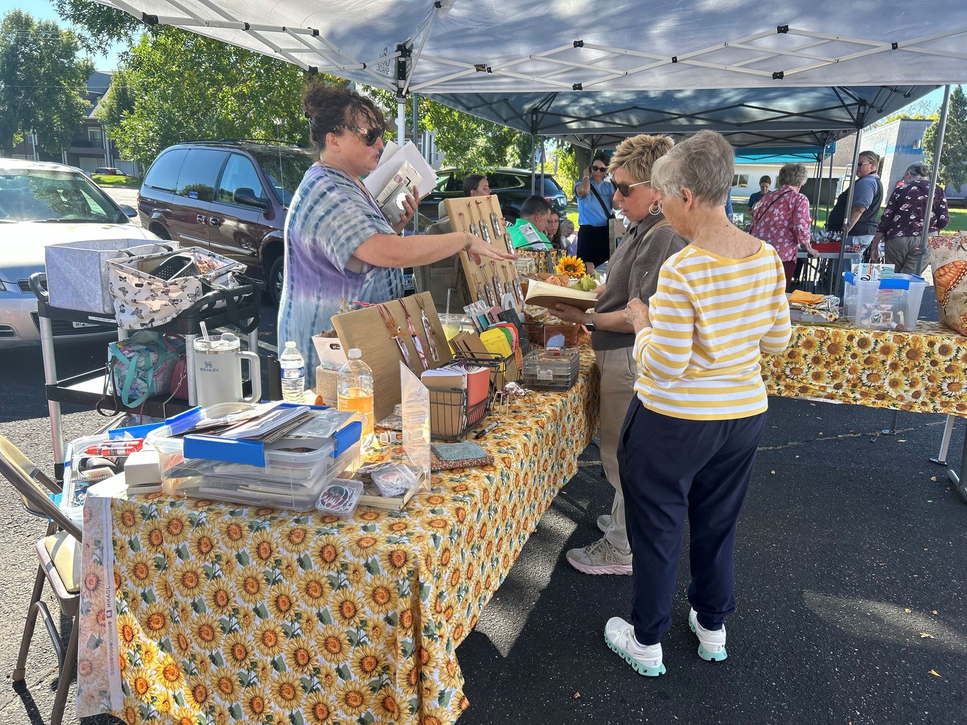 People browse handmade crafts at an outdoor market stall with a sunflower-patterned tablecloth.