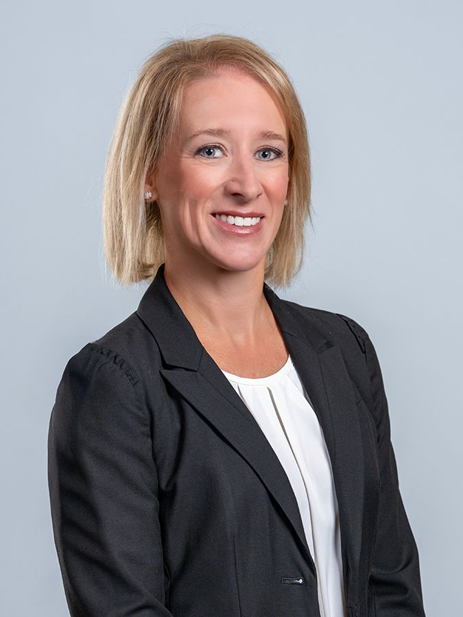 A professional headshot of a person with blonde hair smiling, wearing a dark blazer over a white shirt against a gray.