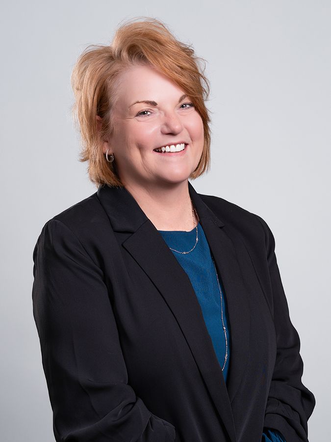 A smiling person with auburn hair wearing a black blazer over a blue top against a light gray background.