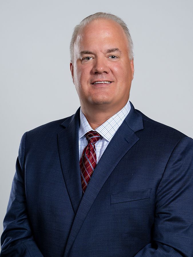 A professional headshot of a person wearing a navy blue suit, a light blue checkered shirt, and a red patterned tie.