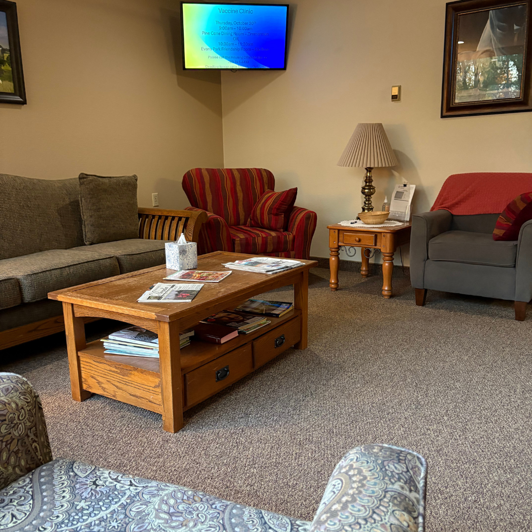 A waiting room with a grey sofa, red patterned armchair, a wood coffee table, a lamp on an end table, and a wall-mounted TV.