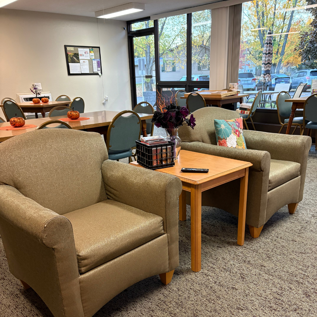 A quiet communal room with tan armchairs, a wooden side table holding a basket and remote, and dining tables in the back.