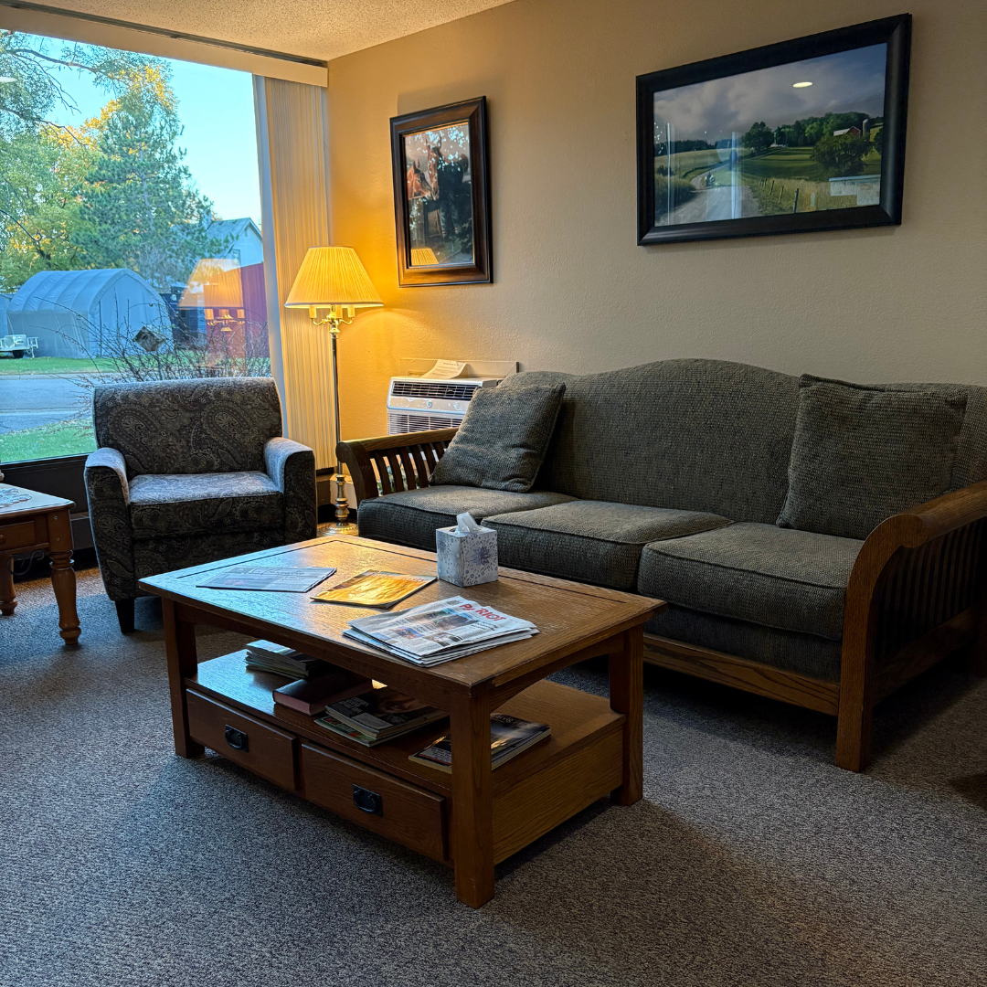 A cozy living room with a green sofa and armchair, a wooden coffee table, and framed art on the wall near a large window.