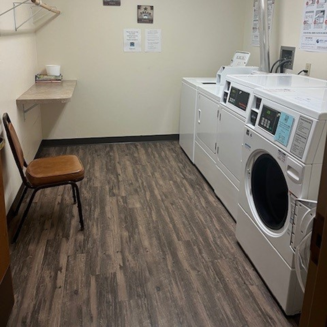 A small laundry room with wood-patterned flooring, a folding table, a brown chair, and two white stacked washer-dryer units.