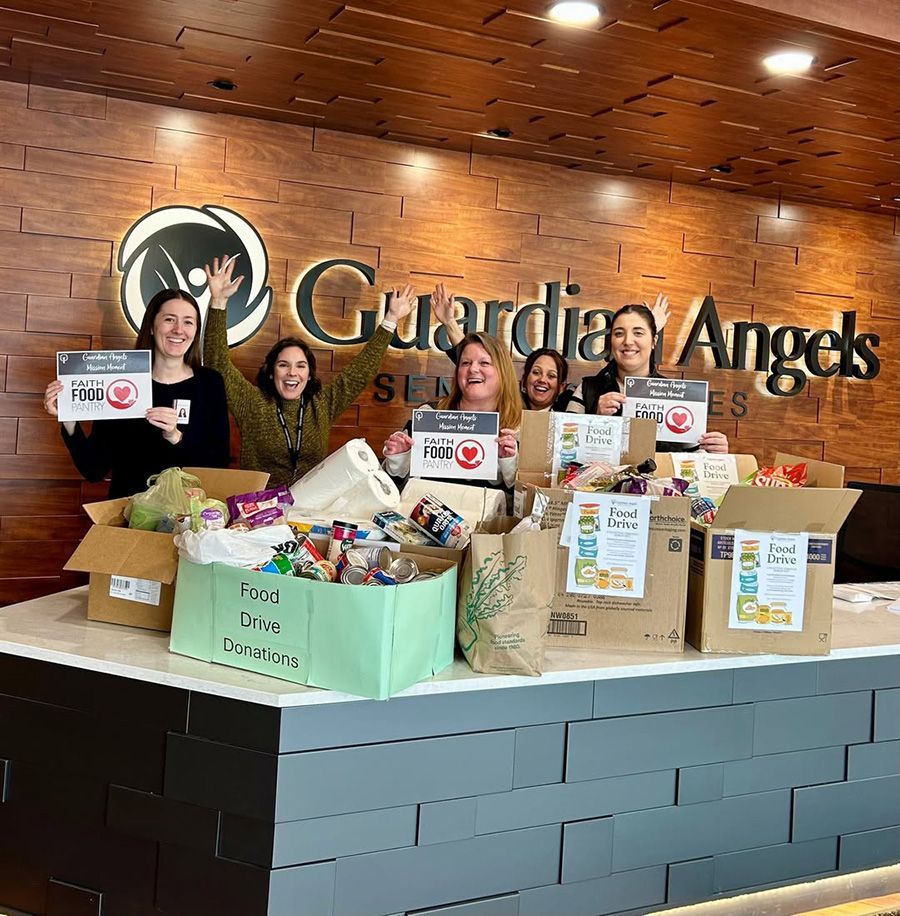 Five people smile behind a counter at Guardian Angels, holding food drive signs behind boxes of donated food.
