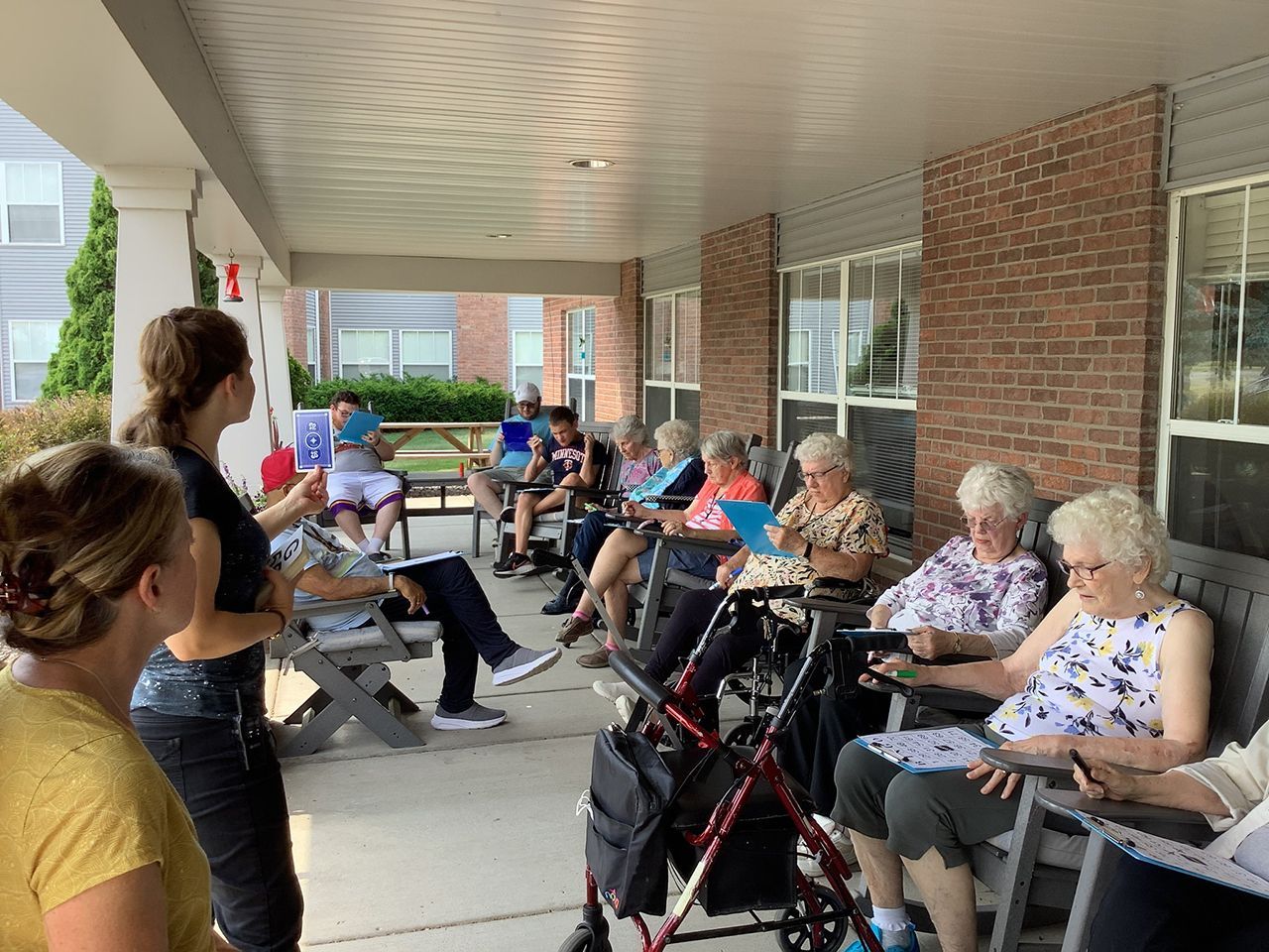 A group of people sit on a shaded porch holding blue activity cards while a leader stands and facilitates.