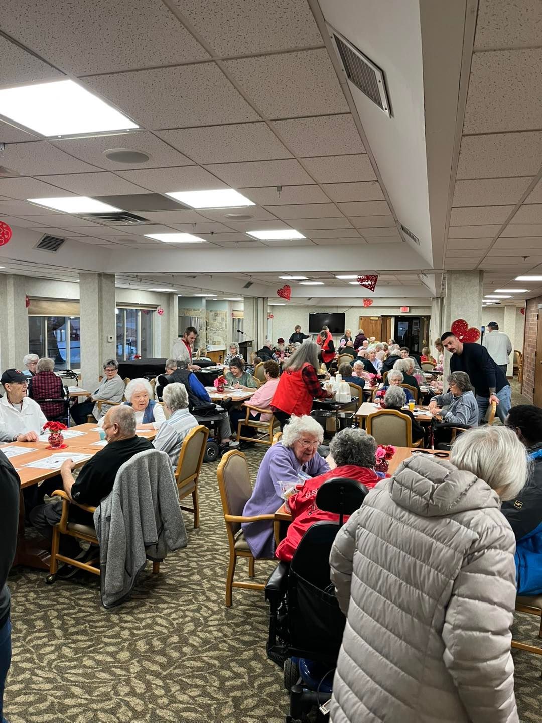 Many people sit at tables in a large, brightly lit dining room decorated with red accents for a social gathering.