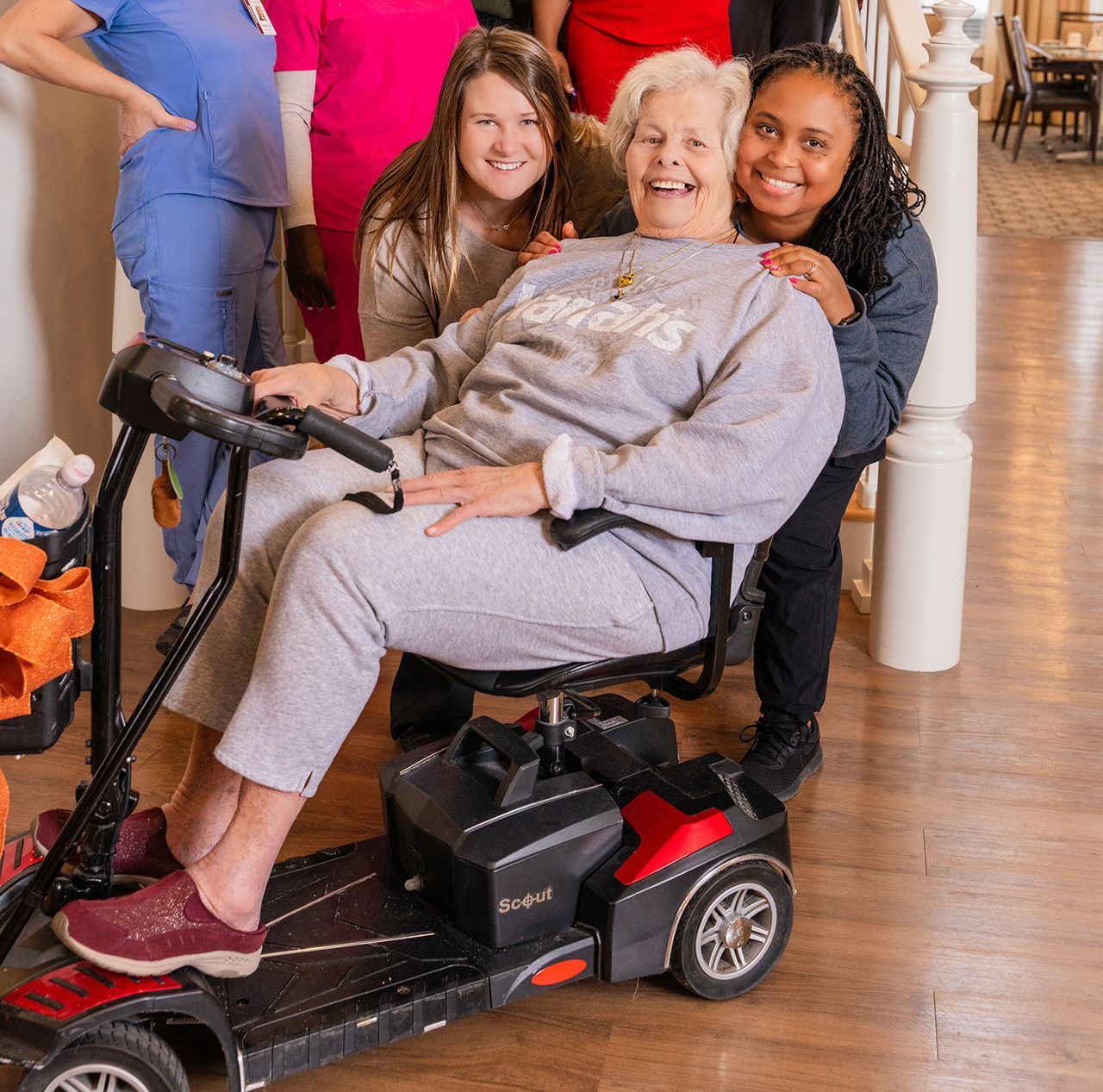 A person in a mobility scooter smiles while two people stand behind, resting their hands on the scooter's seat.