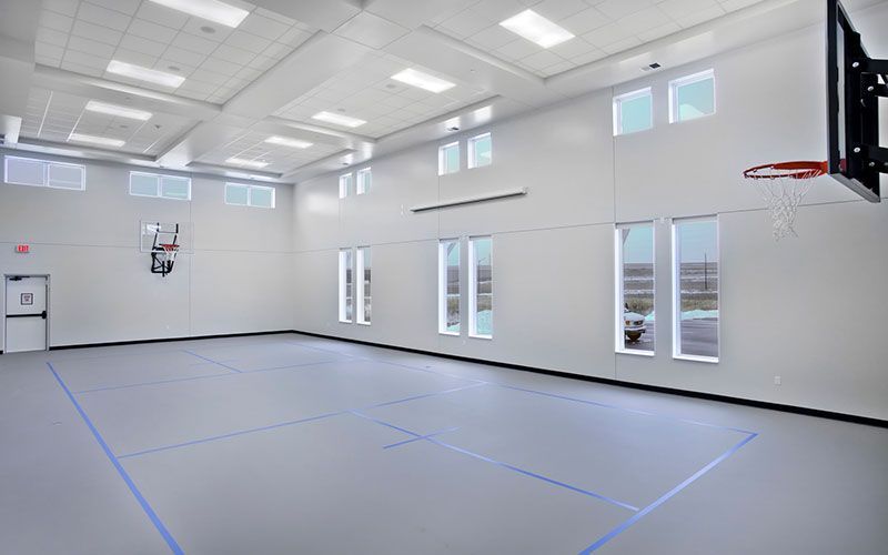 A bright, minimalist indoor gymnasium with white walls, grey flooring, blue court markings, and basketball hoops.
