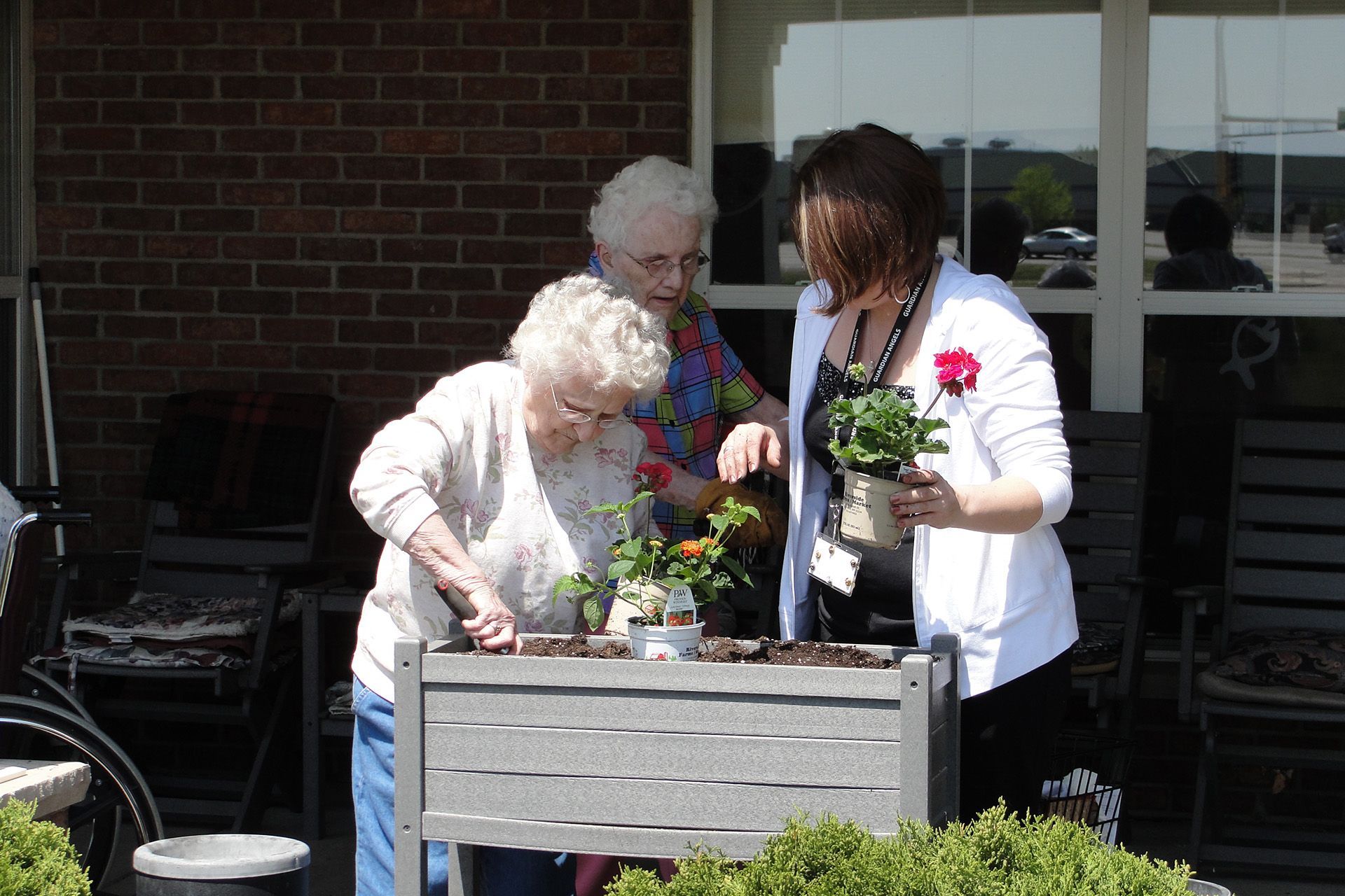 Two people stand at a raised garden bed outdoors, planting flowers together while a third person watches.