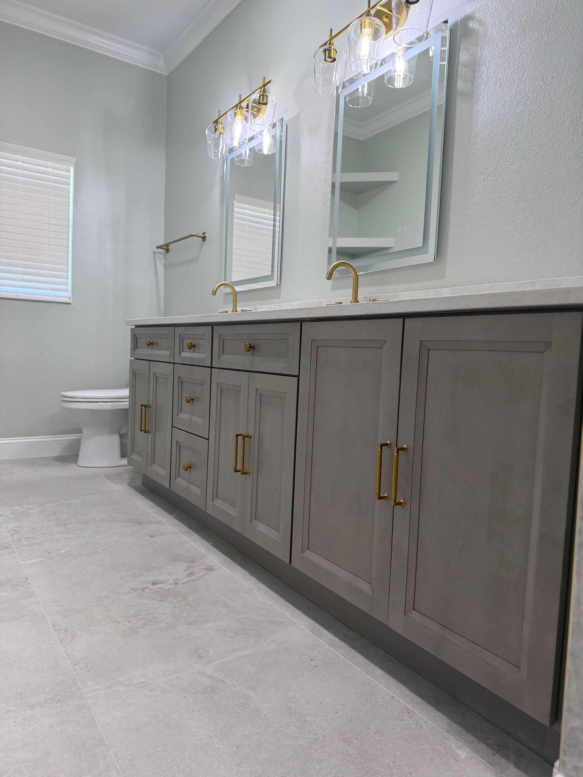 Grey Tile flooring bathroom, with grey shaker cabinets, gold hardware and pull handles, backlit mirrors, and brushed gold lamps.