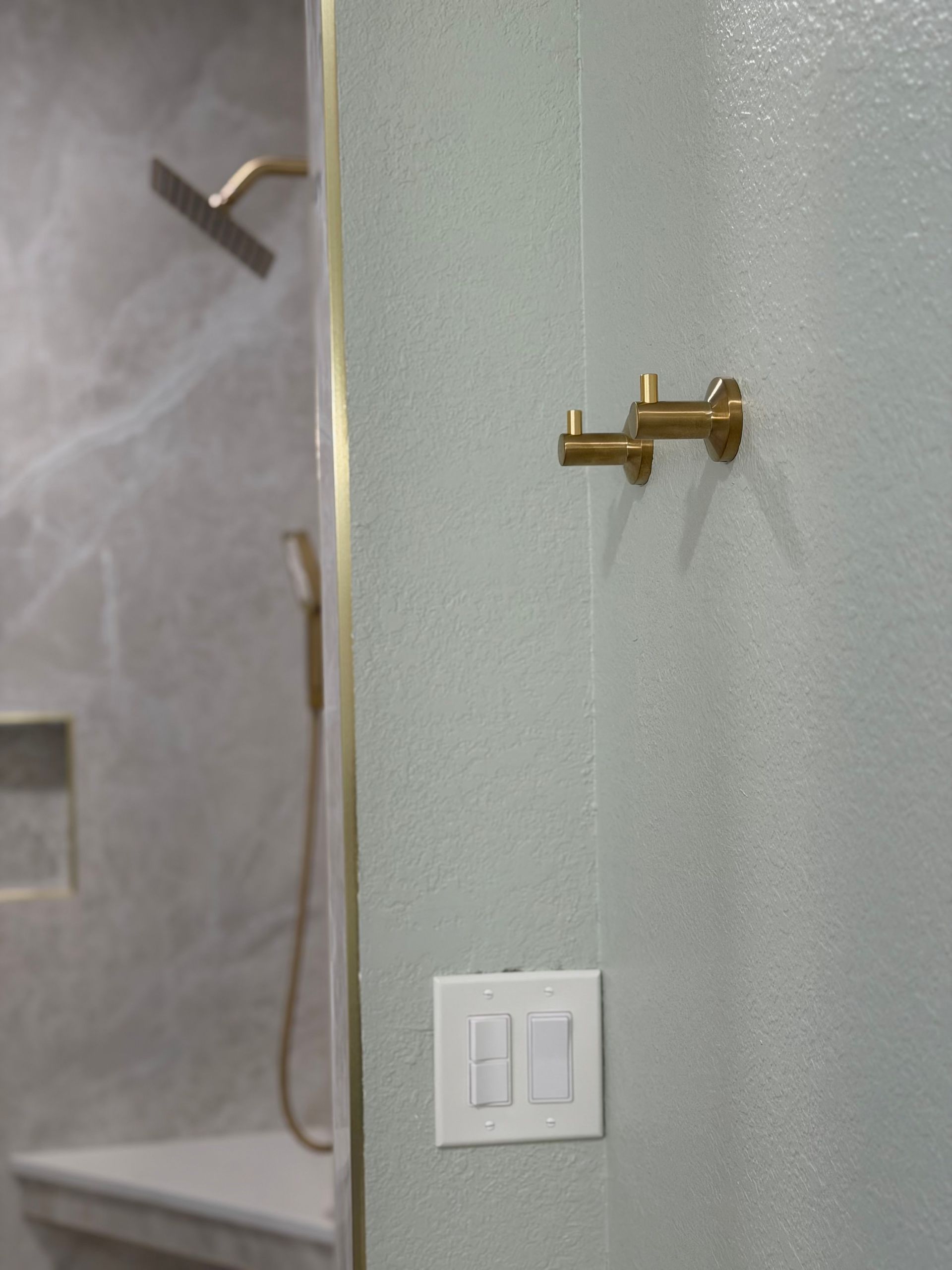 Bathroom shower wall with brass hooks and a white light switch near a glass shower door