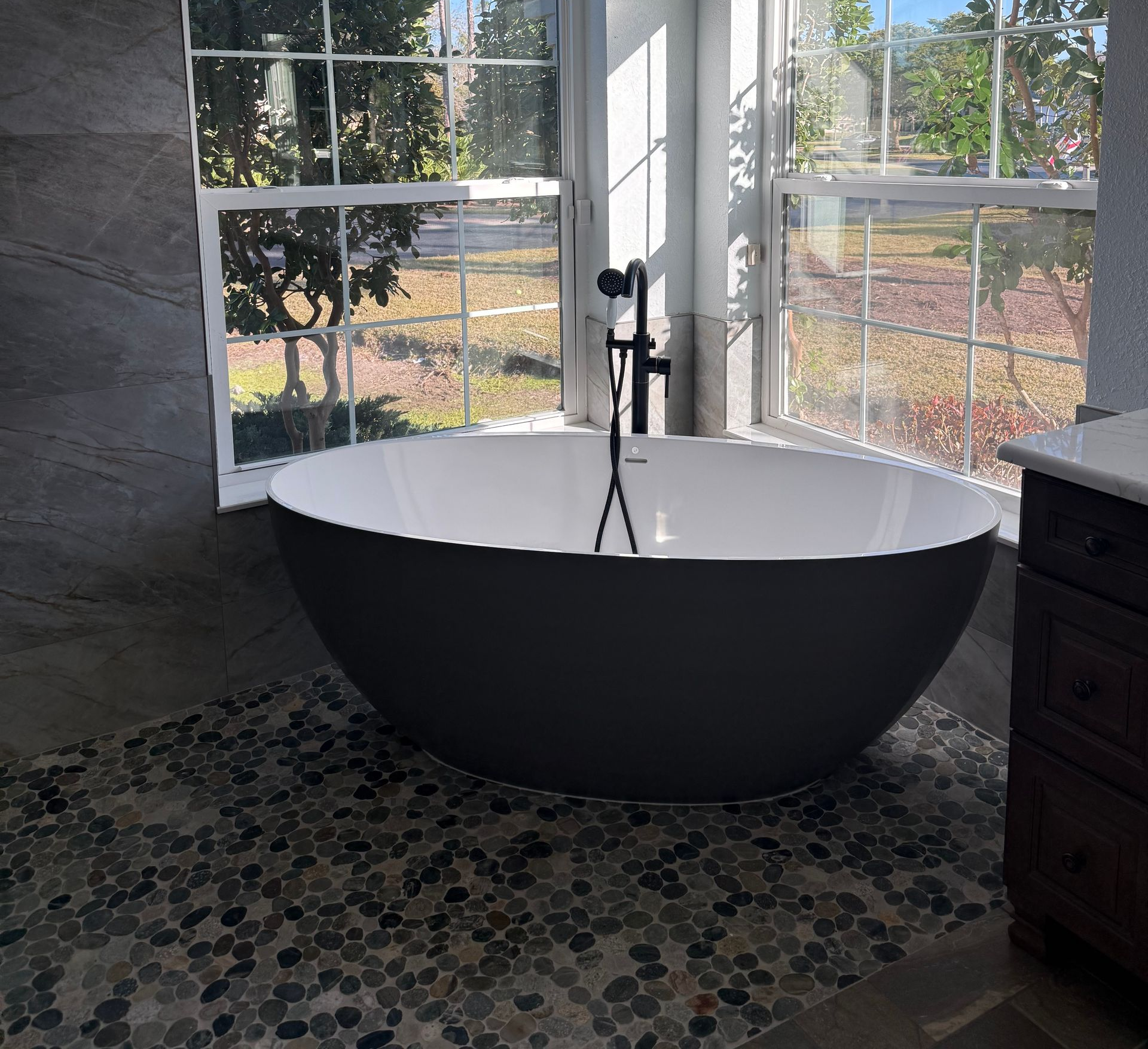 Grey modern tub in bath with matte black trim, two large windows and grey tile walls mosaic floor