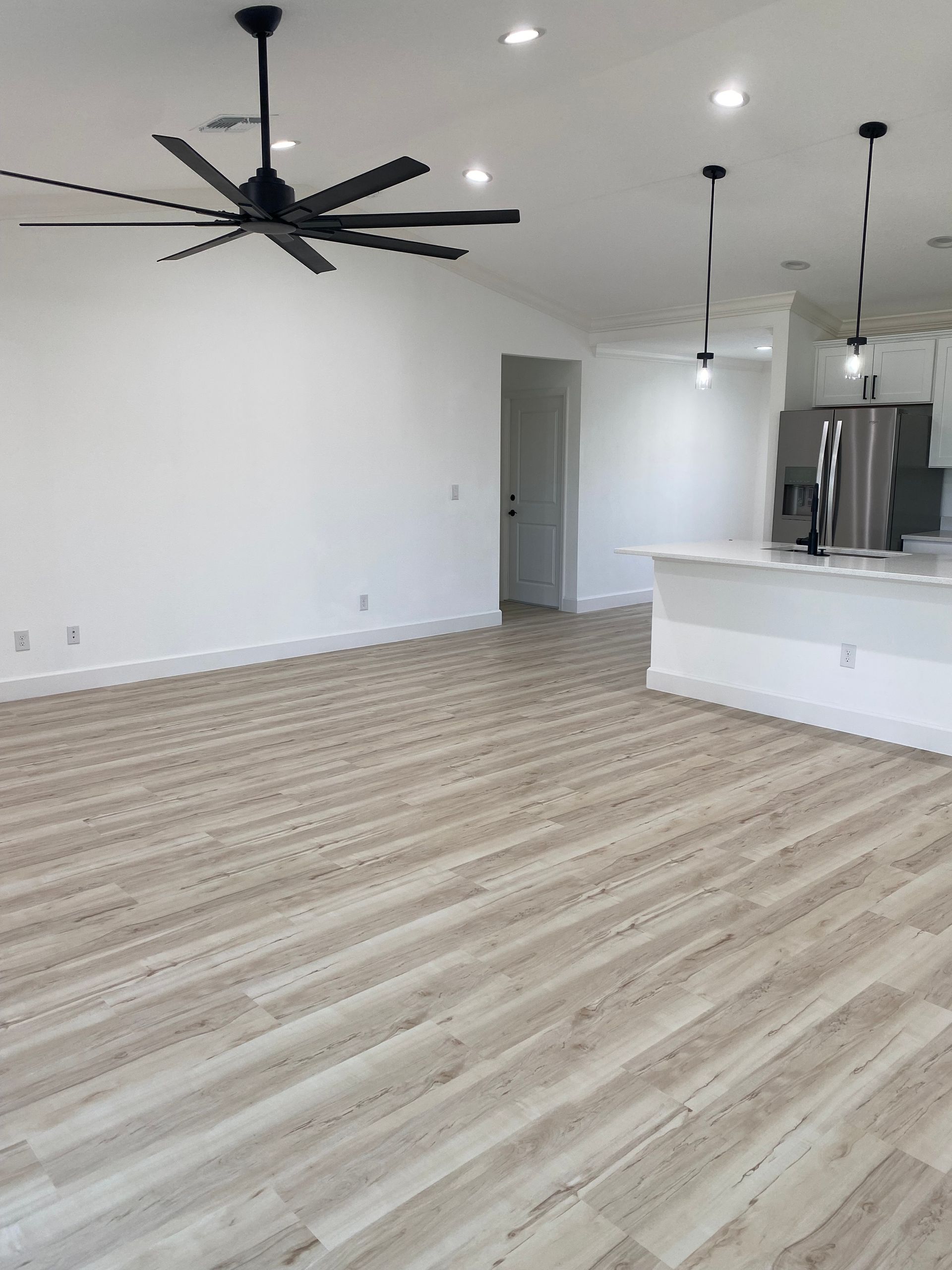 A living room with a ceiling fan and a kitchen in the background.