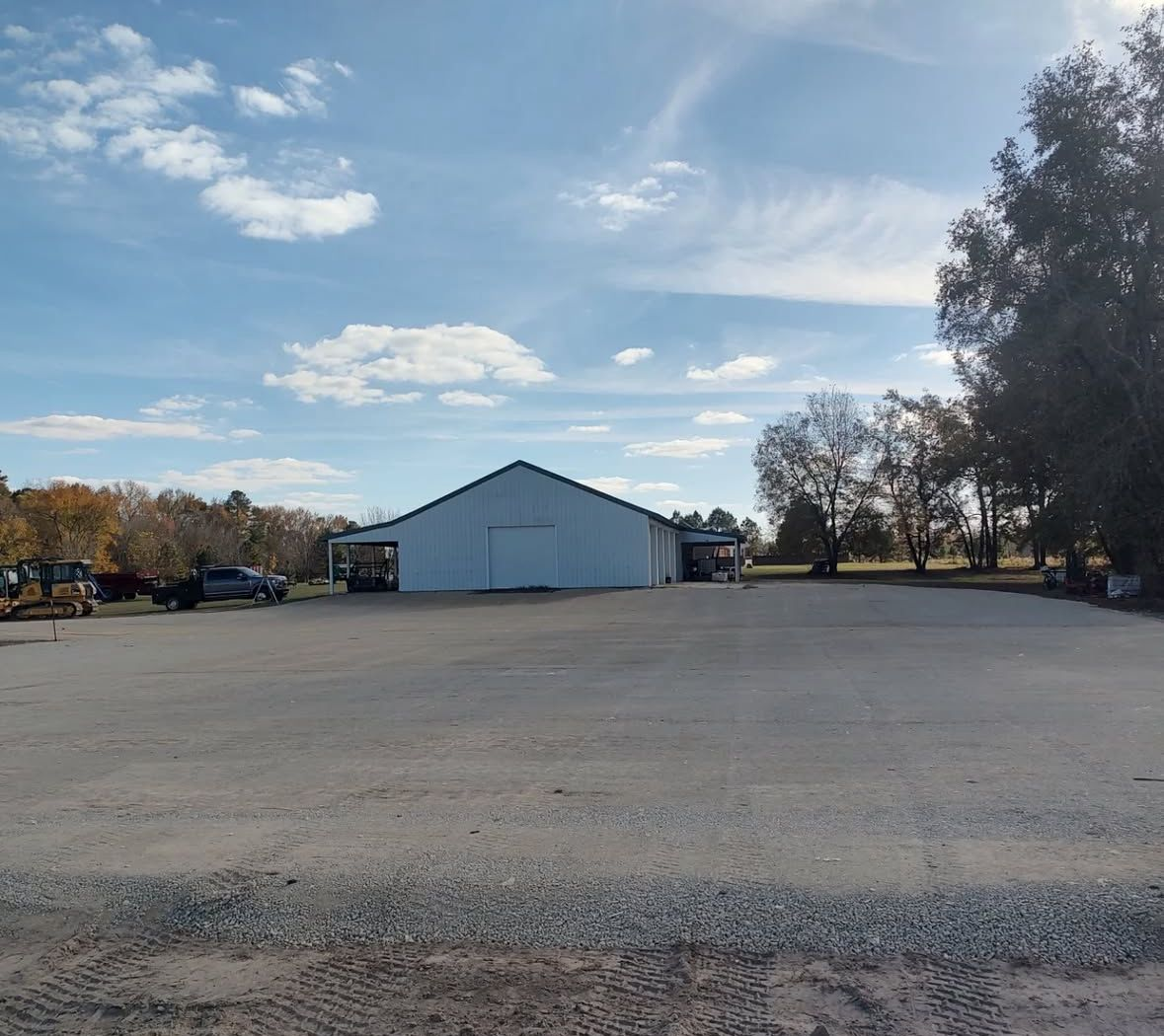 White barn in a gravel lot under a partly cloudy blue sky, with trees in the background.