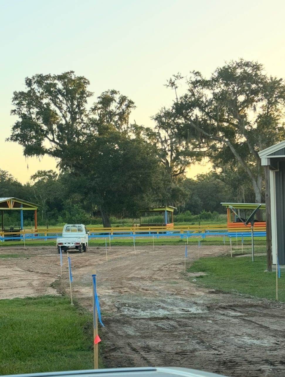 A white truck drives on a dirt road, trees and colorful structures in the background, daytime.