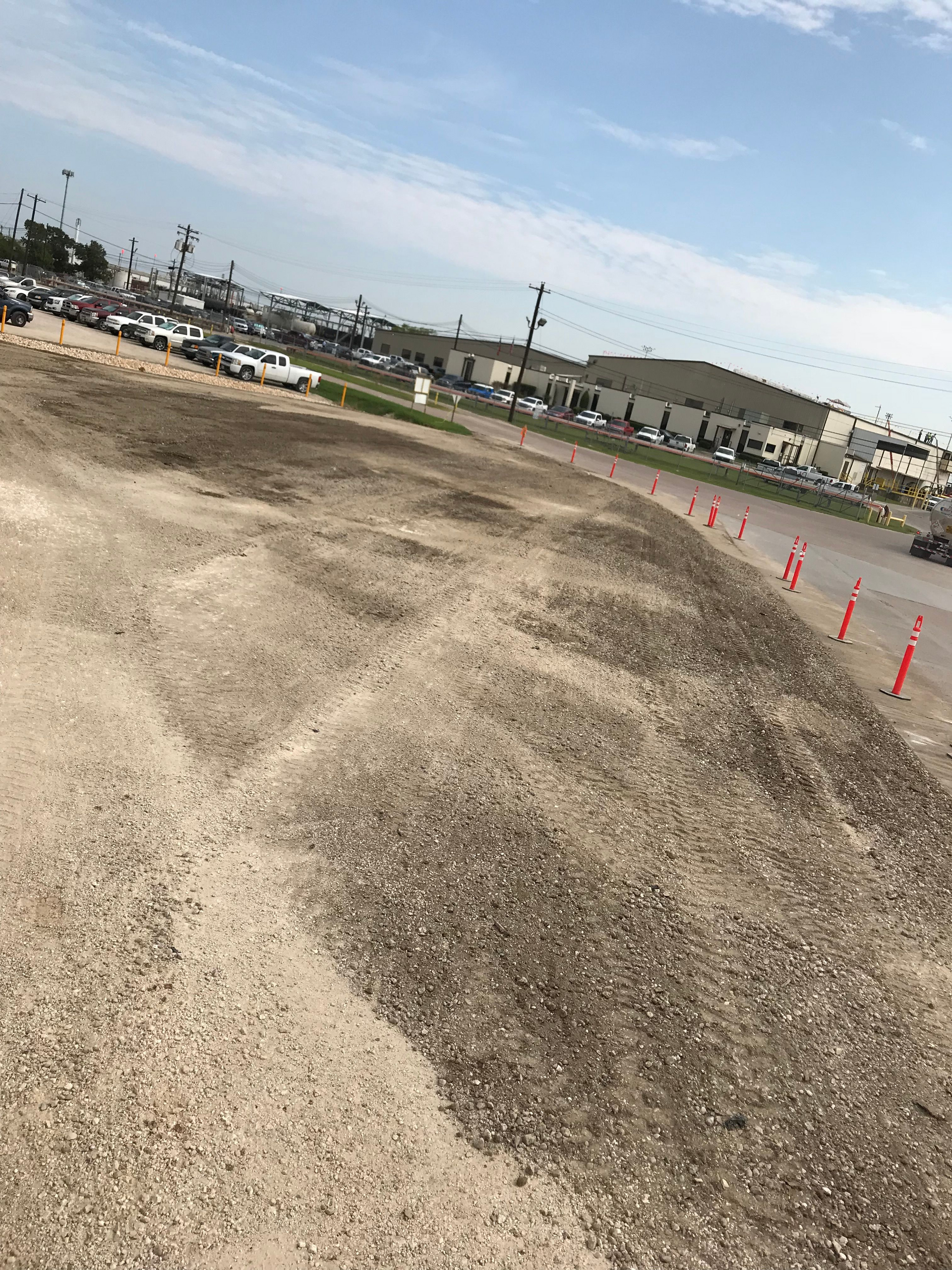 Gravel pile in open lot with cars, buildings, and sky in the background.