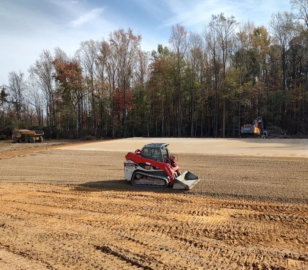 A red and white skid steer grading soil at a construction site, trees in the background.