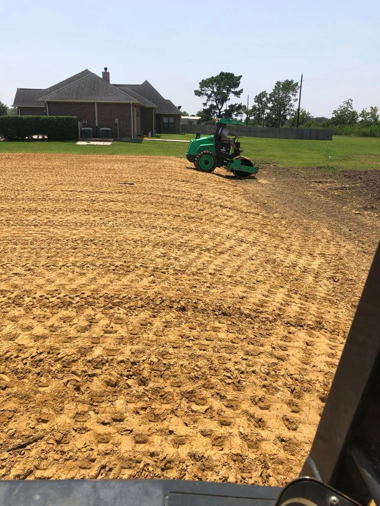 A green lawn roller compacts soil in an open field, near a house on a sunny day.