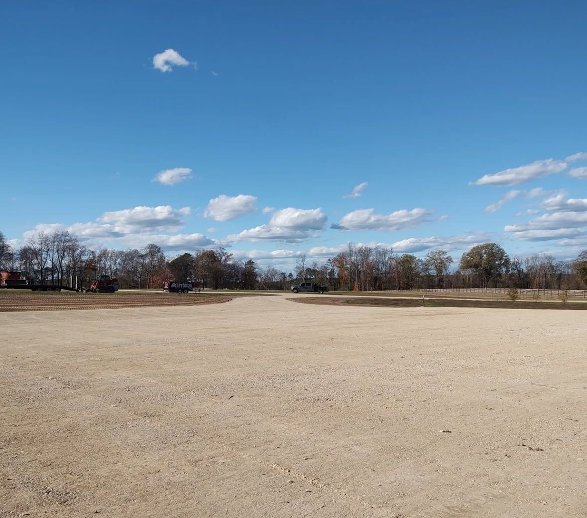 A wide, empty, gravel lot under a bright blue sky with scattered clouds and a tree line.