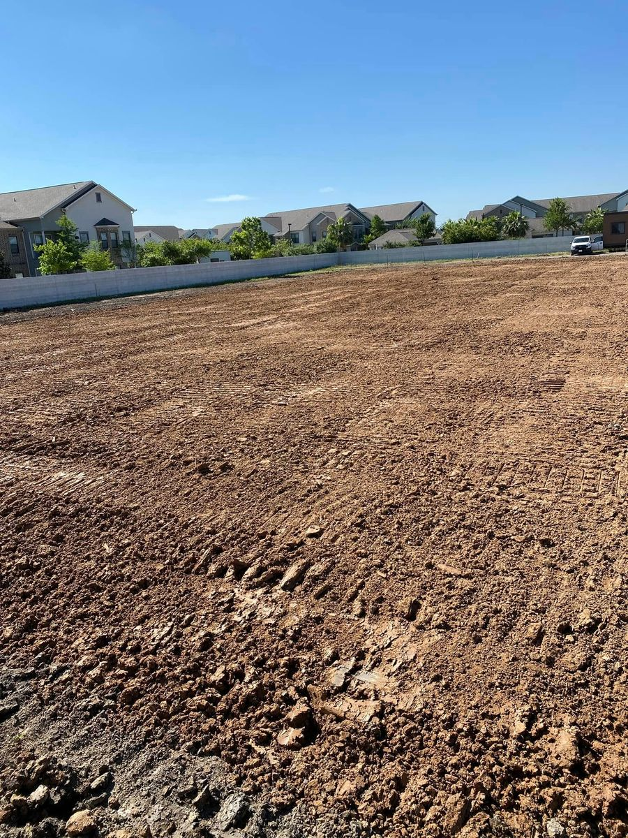 Cleared lot of brown dirt with tire tracks, houses and a white fence in the background under a blue sky.