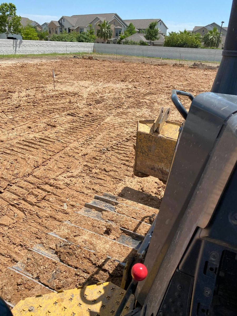 View from inside a bulldozer cab, looking at a freshly tilled dirt field on a sunny day. Houses are in the background.
