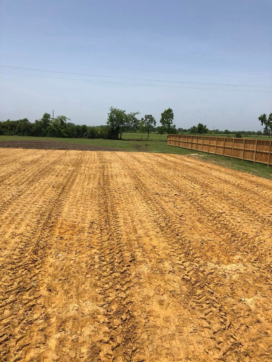 Field of tilled, brown earth with tire tracks, bordered by a wooden fence, green grass, and trees under a blue sky.