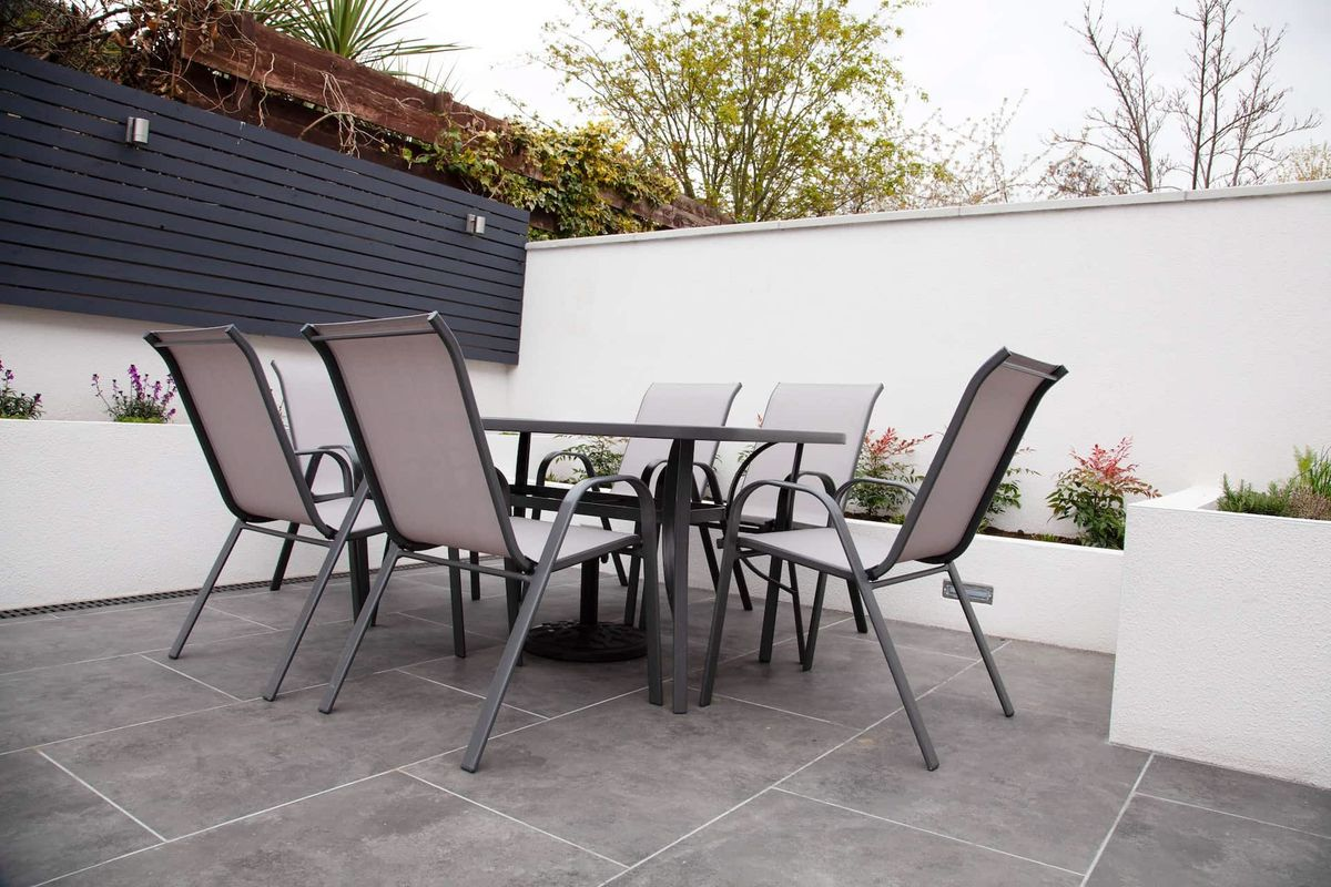 Patio with grey dining set on stone tiles, surrounded by white walls and black fence, with plants.