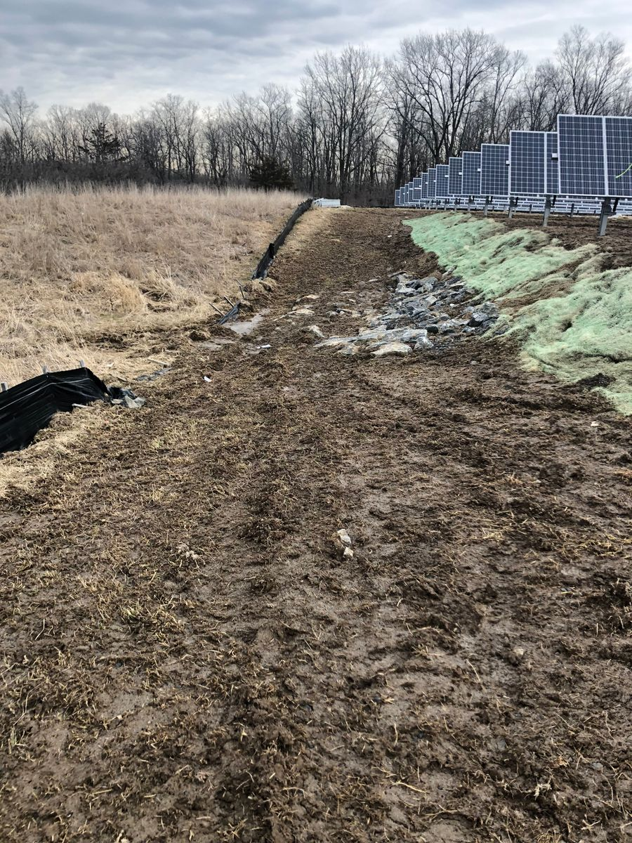 A muddy, eroded ditch next to a solar panel array and a field of dry grass under a cloudy sky.
