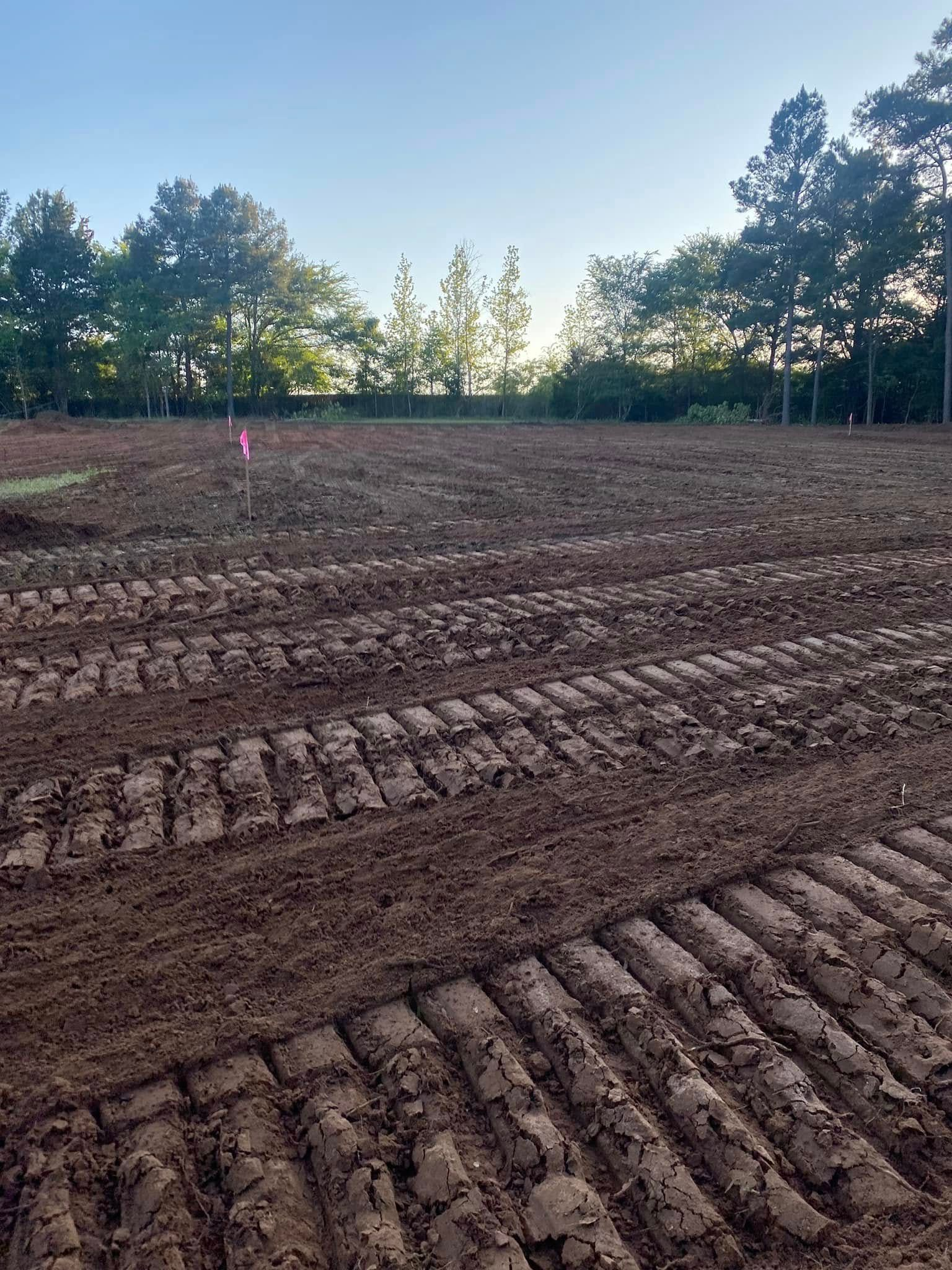 Tilled field with tire tracks, pink flags, and trees in background.