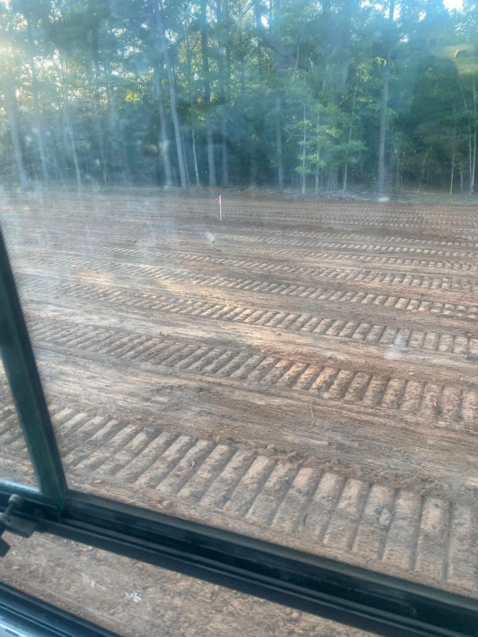 View from a vehicle's window of a recently graded dirt plot with tire tracks, trees in the background.
