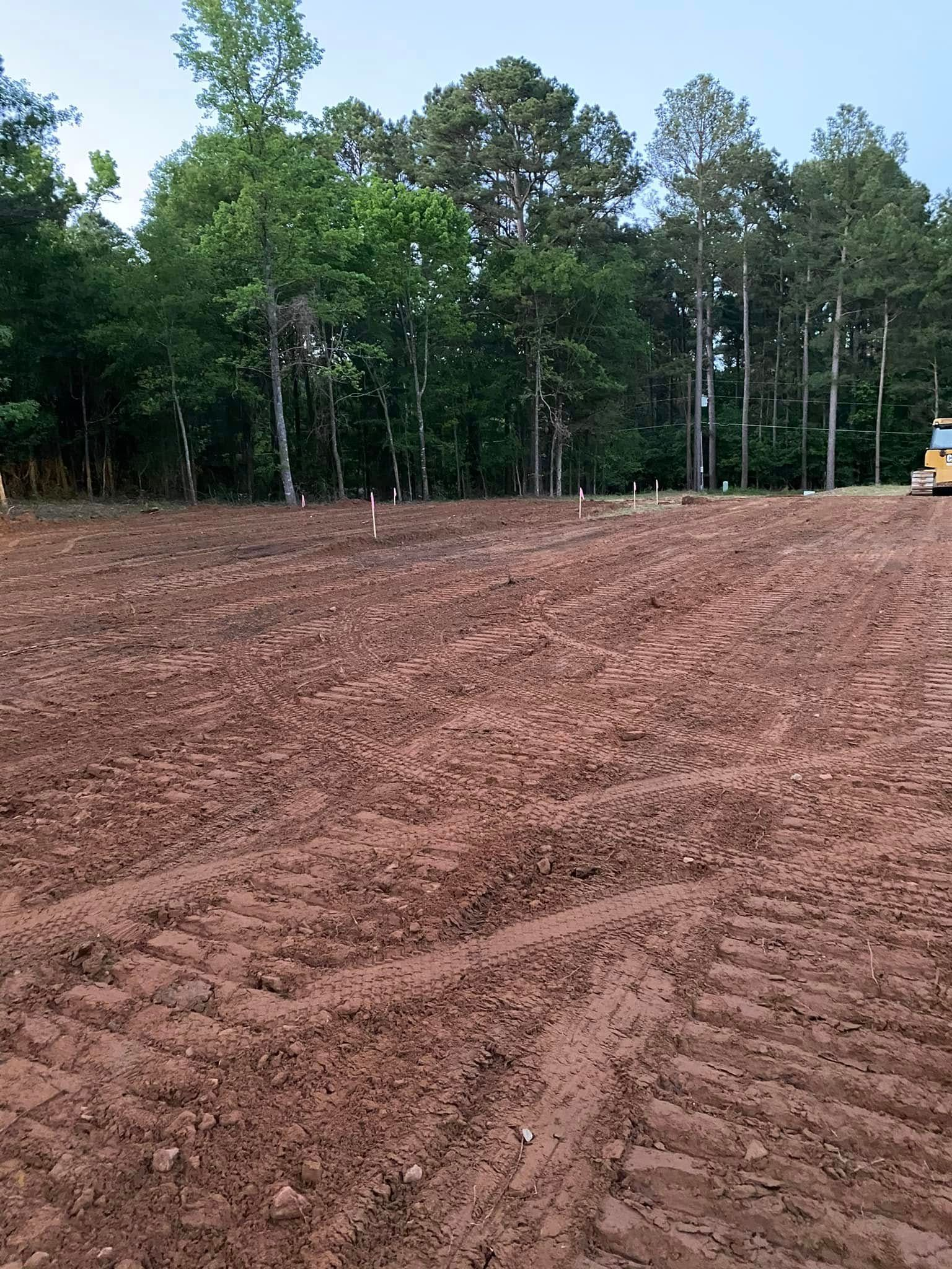Cleared dirt lot with tire tracks, small markers, and a tree line in the background under a blue sky.