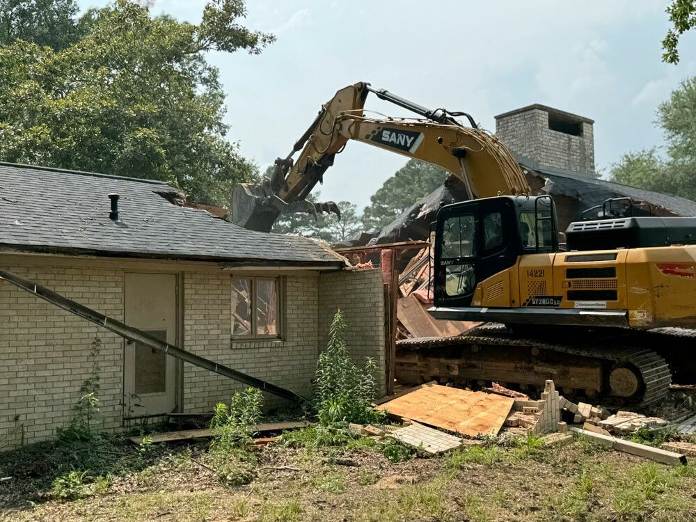 Excavator demolishing a brick house; the roof and walls are being torn down.