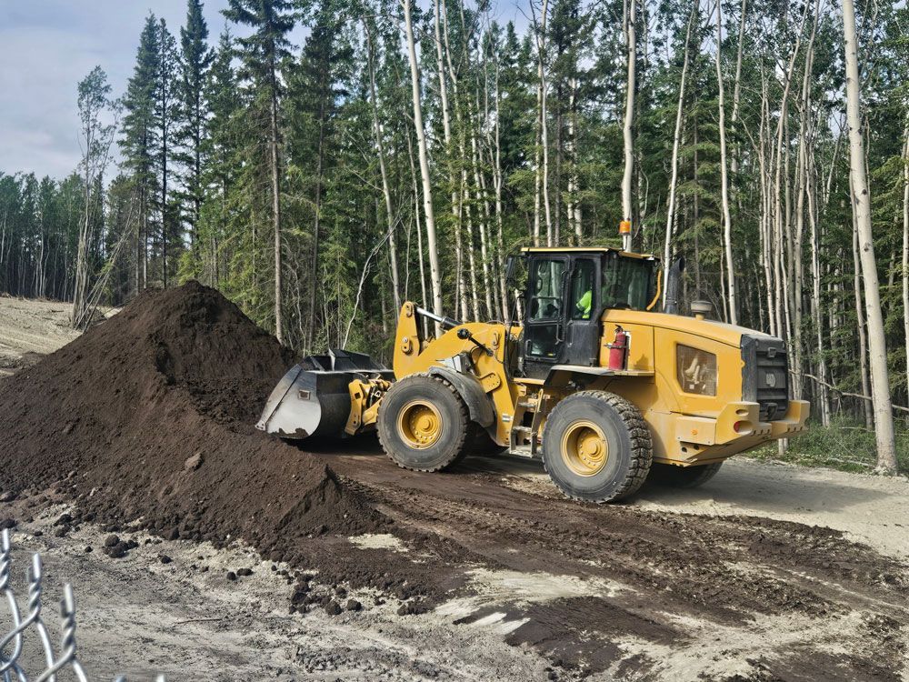 Yellow front loader moving dirt on a forest road beside a large soil pile.