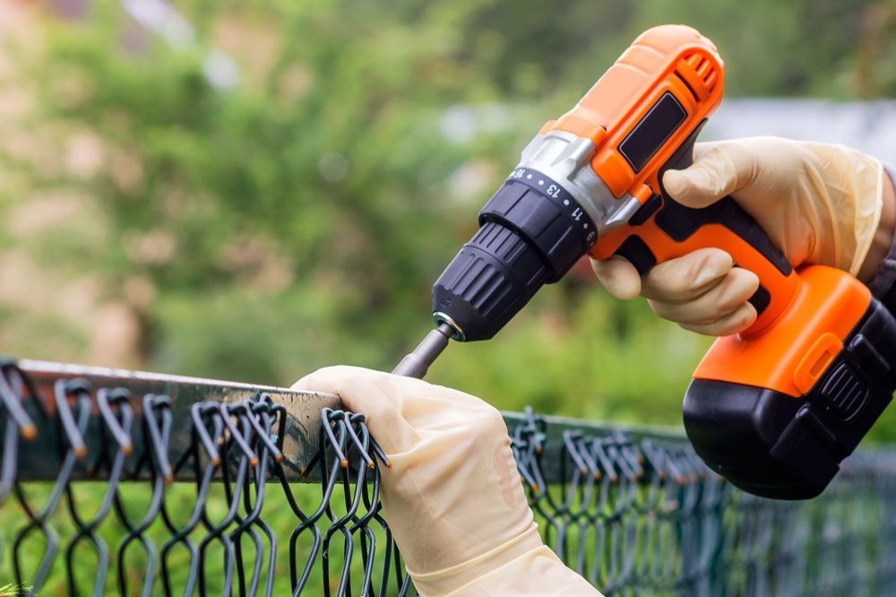 Gloved hands using a drill to install a black chain-link fence panel outdoors