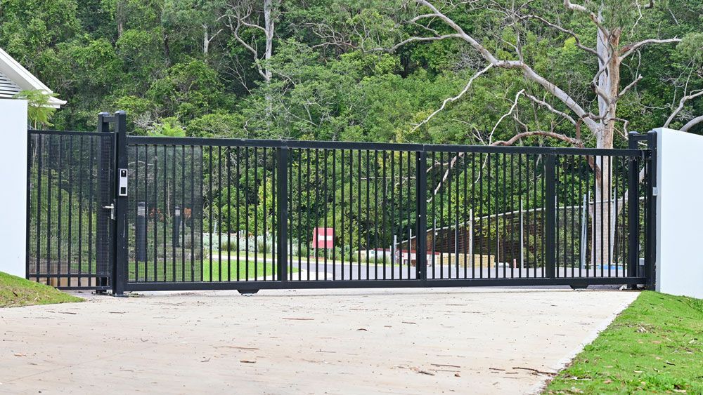 Black metal sliding gate blocking a driveway, with trees and greenery behind it.