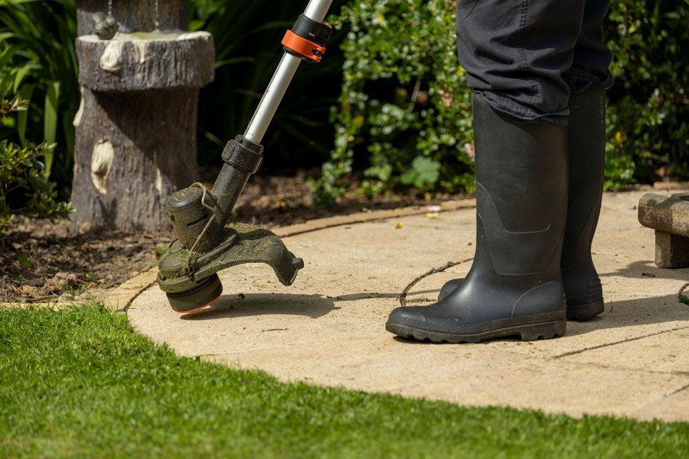 Person using a string trimmer to cut grass along a garden path, wearing black boots.