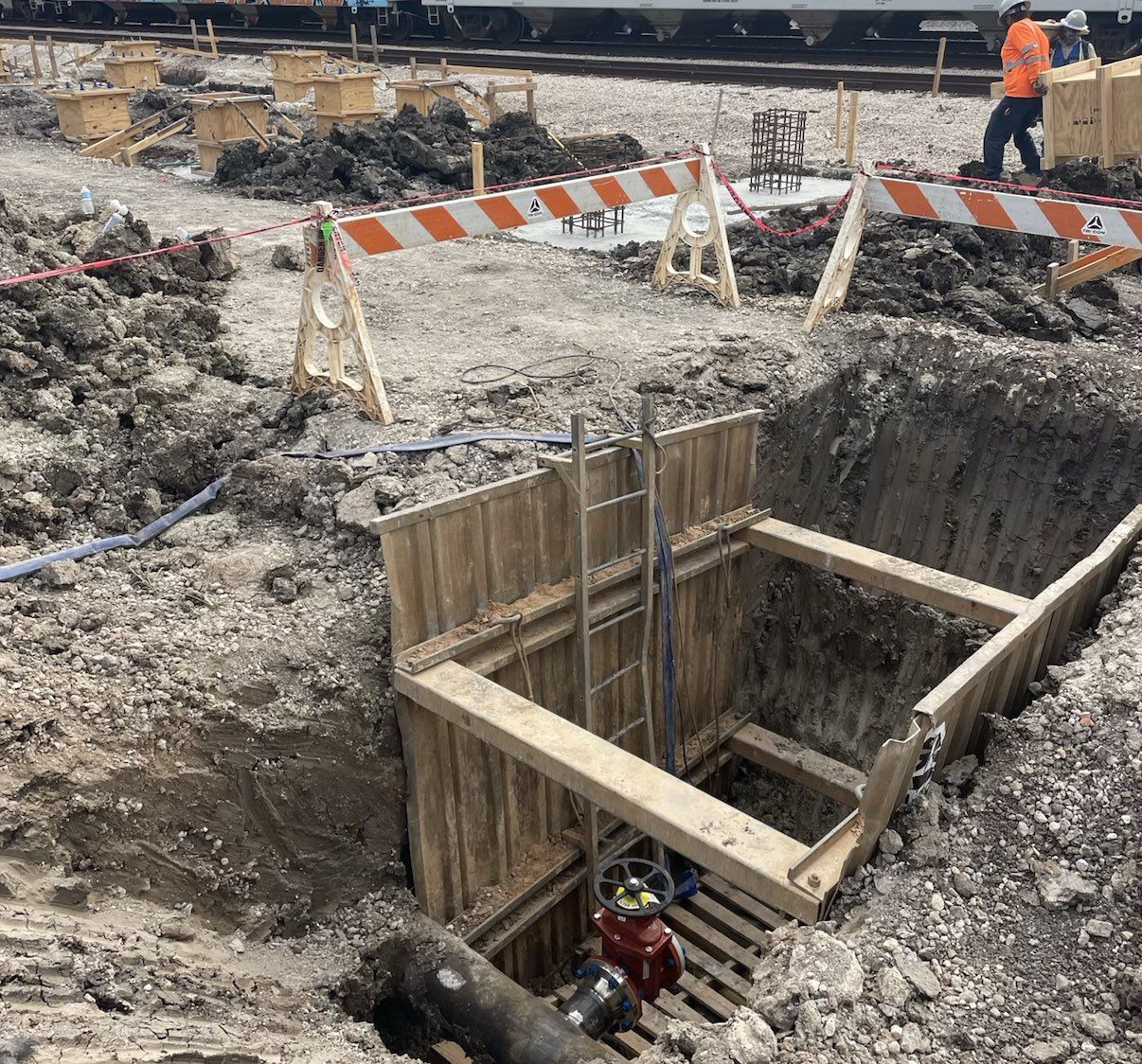 Construction site with wooden trench, ladder, barriers, and worker near train.