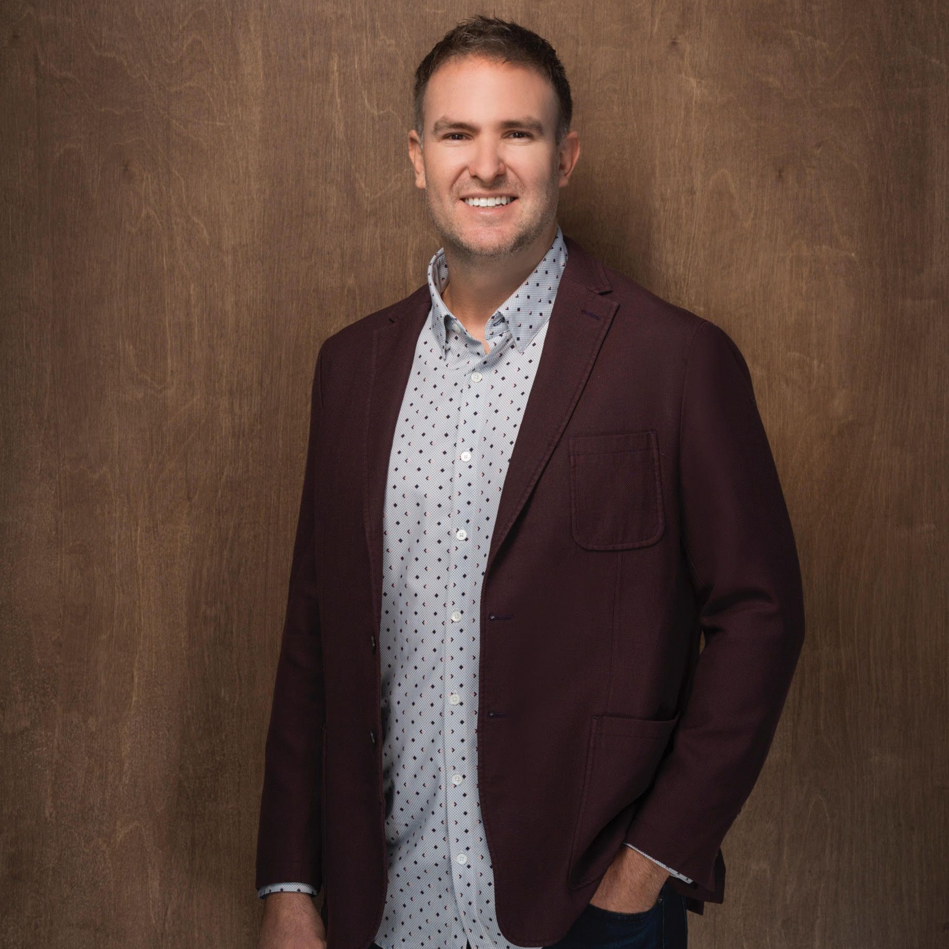 Man standing in a burgundy blazer and patterned shirt against a brown studio backdrop