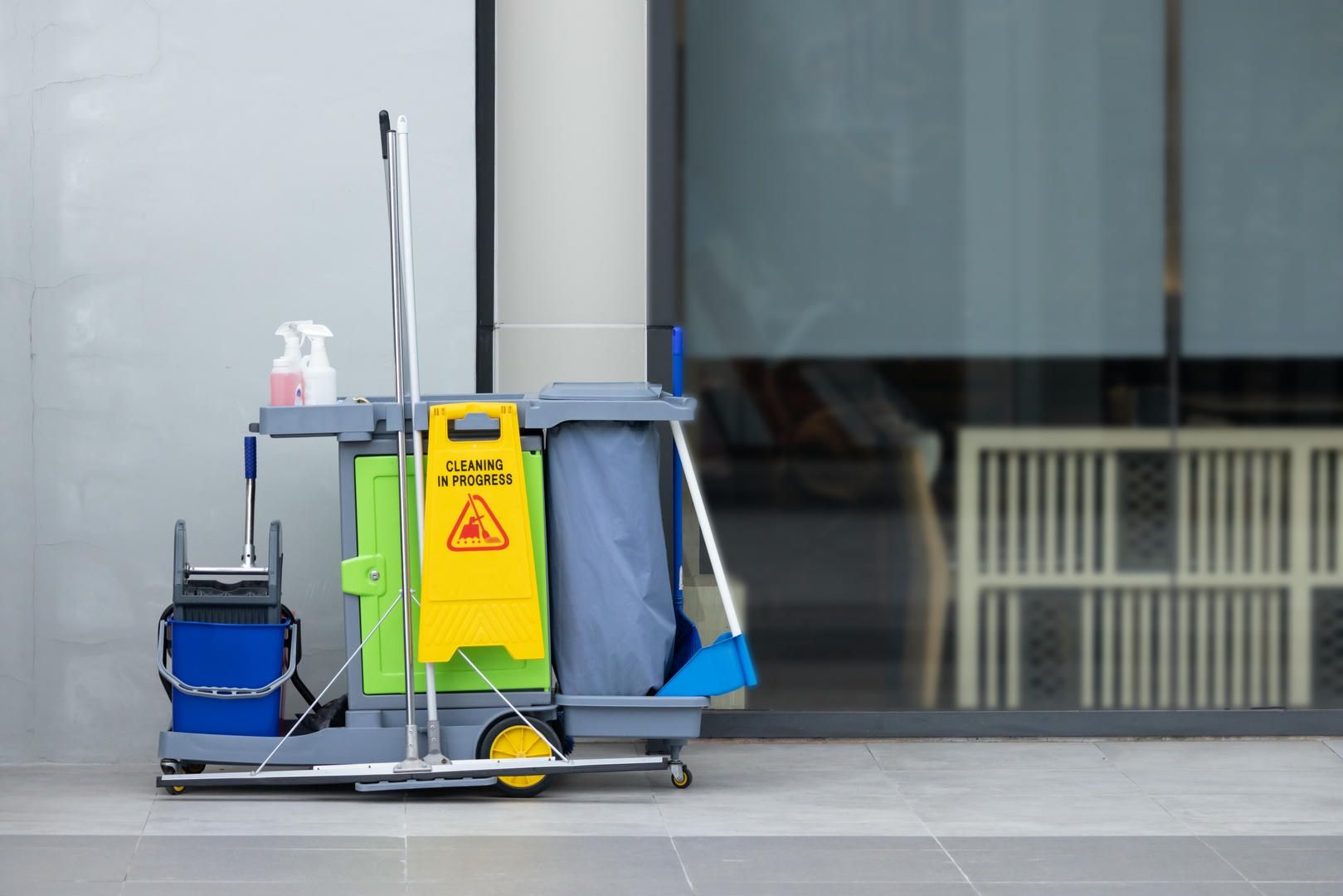 A cleaning cart with a yellow caution sign on it is parked on the sidewalk in front of a building.