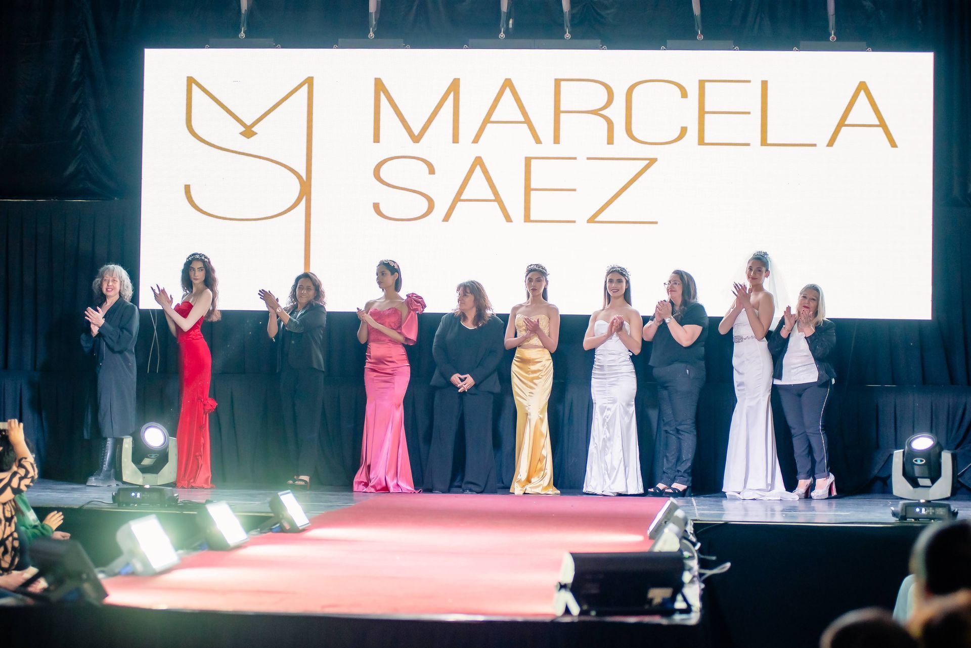 Desfile con modelos posando bajo un brillante telón de fondo de “Marcela Saez”.